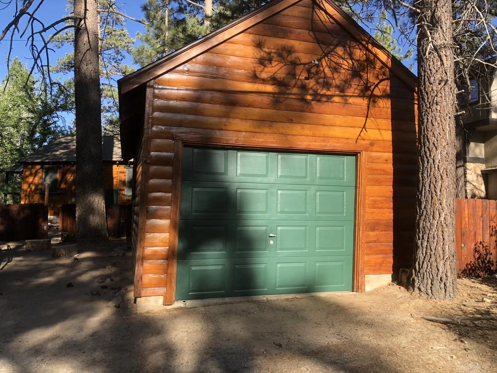Freshly painted cabin garage in Big Bear, warm stained log siding and a clean forest-green door surrounded by pines.